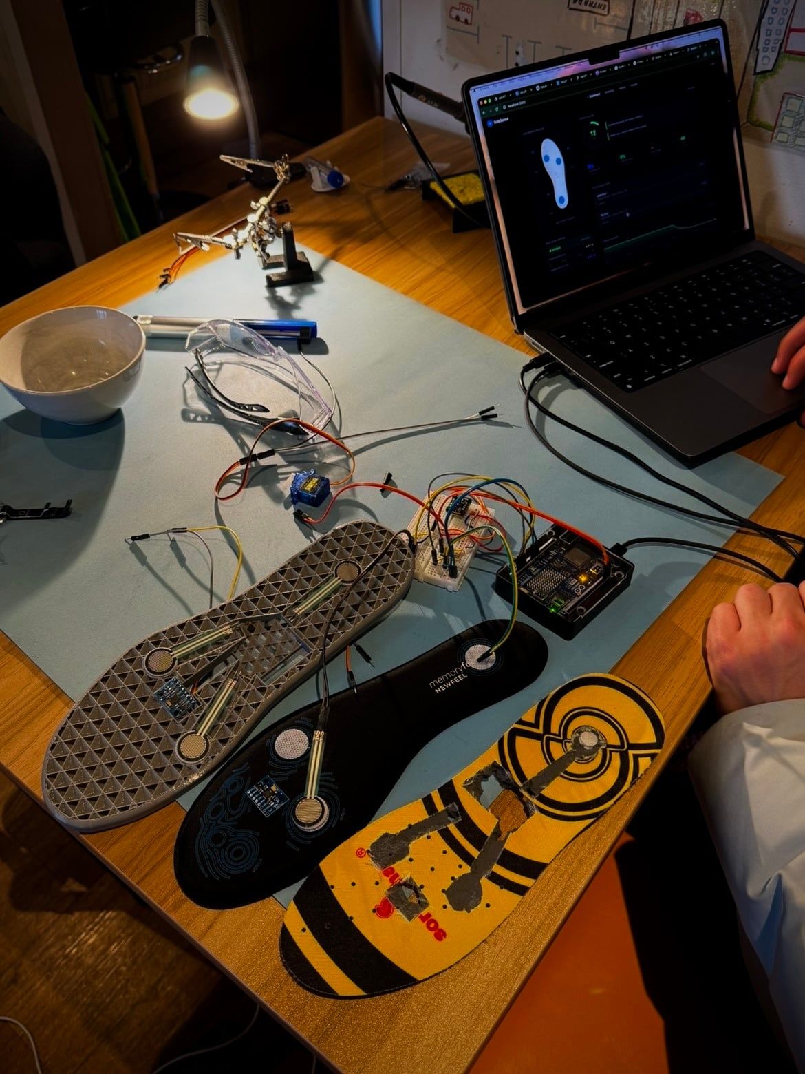 The SoleSense prototype on a workbench: three insoles carrying force sensors, wired through a breadboard to an Arduino, with the live dashboard open on a laptop.